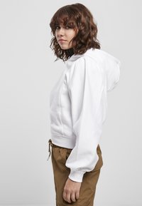 Hoodie bianco cropped con cappuccio regolabile, polsini a coste e vestibilità comoda, abbinato a pantaloni marroni a vestibilità rilassata.