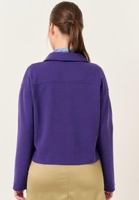 Paarse cropped jack met een kraag en lange mouwen. Zachte stof, naadloos ontwerp en rechte snit aan de achterkant. Gecombineerd met een beige rok.