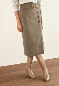 Jupe crayon à carreaux marron avec une taille ceinturée et cinq boutons sur le côté, assortie à des bottes à talons beiges jusqu'aux genoux sur un sol en bois.