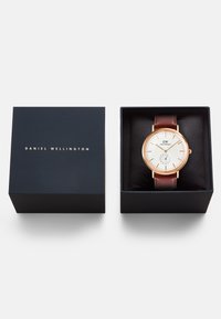 Daniel Wellington laikrodis tamsiai mėlyname dėkle. Savybės: baltas ciferblatas, rožinio aukso korpusas, mažas subciferblatas ir rudas odinis dirželis.