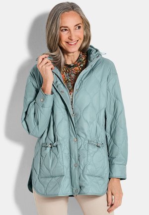 Frau mit grauen Haaren, die eine hellblaue gesteppte Jacke über einem gemusterten Oberteil trägt, lächelt und hält den Jackenkragen mit einer Hand.