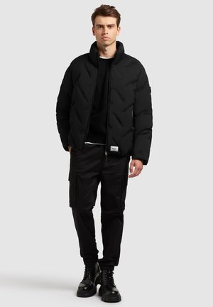 Sort dunjakke med quiltet design, høj krave og lynlås, båret over en sort sweatshirt og sorte cargo-bukser, parret med sorte støvler.