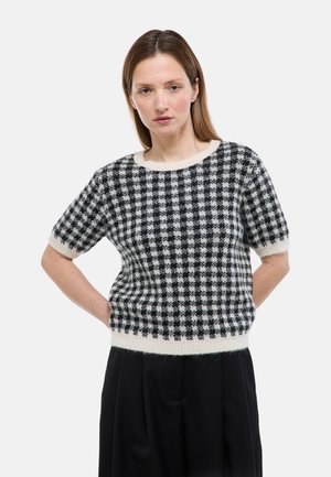 Femme aux cheveux bruns raides porte un pull à manches courtes à carreaux noirs et blancs et une jupe noire, debout les mains sur les hanches.