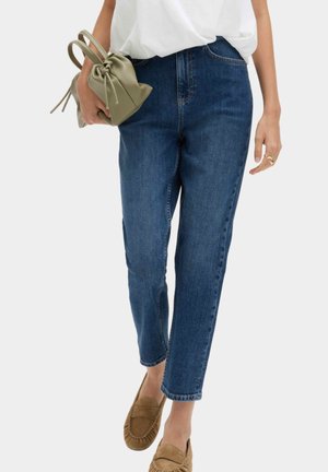 Kvinde iført blå jeans med lige ben, hvid top, brune loafers, der holder en lille olivengrøn taske med snøre i venstre hånd.