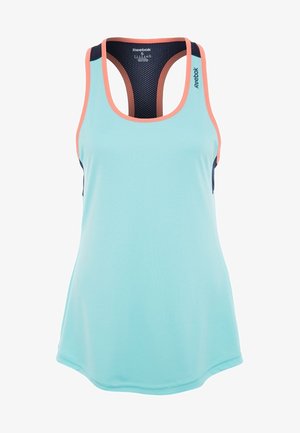 Femme portant un haut de sport turquoise sans manches avec une bordure corail, des leggings courts noirs et des chaussures de sport noires, debout devant un fond uni.