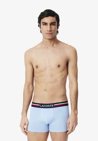 Boxers légers bleu clair en matériau doux avec une taille rayée navy et rouge portant le logo Lacoste. Le modèle se tient contre un fond uni.