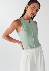Top senza maniche verde menta con zip frontale, caratterizzato da un fit sartoriale e linee pulite. Abbinato a pantaloni bianchi a vita alta.