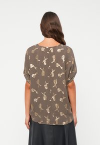 Brun bluse med gullmønstrede detaljer; myk stoff, løs passform, korte ermer, avrundet nederkant, minimalistisk design; vist fra bak.