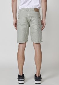 Vyras, vilkintis šviesiai pilkas sukurtas džinsinis šortai, sukabinti iki kelių, ir juodas Koröshi sportinis batų, stovi atsukęs nugarą prieš pilką fono.