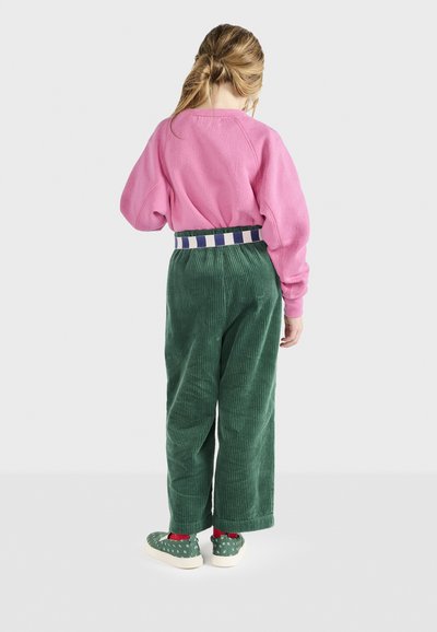 Rosa Sweatshirt mit abgesenkten Schultern, grüne breiten Hosen aus Cord, gestreifter Gürtel und gemusterte grüne Schuhe mit roten Socken.