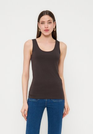 Mladá žena s dlhými tmavými vlasmi, oblečená v čiernom tielku bez rukávov a modrých džínsoch, stojaca pred jednoduchým bielym pozadím.