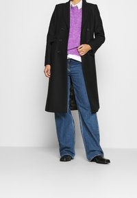 Black double-breasted coat over a purple knit sweater, white shirt, and wide-leg blue jeans. Black boots complete the outfit.
