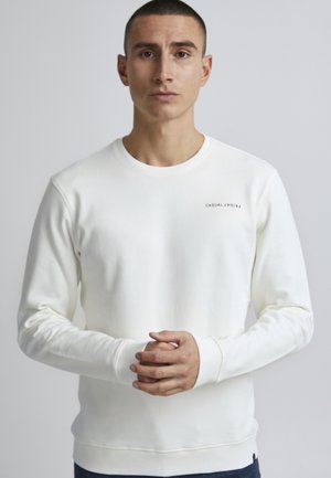 White sweatshirt with long sleeves, round neck, ribbed cuffs and hem, featuring a small text logo on the left chest area. Soft texture.