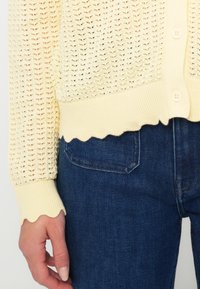 Κίτρινη πλεκτή ζακέτα με λοξό τελείωμα και μανίκια, διακοσμημένη με υφασμάτινο σχέδιο, συνδυασμένη με σκούρο μπλε τζιν. Ορατό κλείσιμο με κουμπιά.