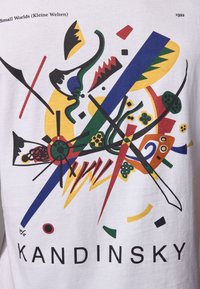 Valkoinen T-paita, jossa on värikäs abstrakti printti Kandinskylta, rohkeilla muodoilla sinisen, keltaisen, punaisen, vihreän ja mustan sävyissä. Alapuolella teksti "KANDINSKY."