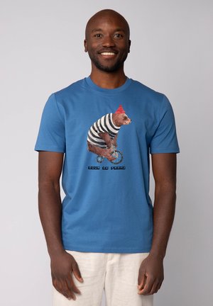 Blauwe katoenen t-shirt met een afbeelding van een beer in een rode hoed en gestreept shirt, die op een eenwieler rijdt met de tekst "YOU’RE BE FRANZ" eronder.