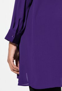 Fialová blůza s dlouhými rukávy, která má zapínání na knoflíky na manžetách. Tkanina je hladká s volným střihem a má mírný lesk.