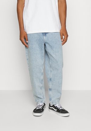 Personne portant un jean ample bleu clair, des baskets noires et blanches, et un T-shirt blanc uni, debout devant un fond uni.