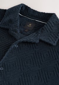 Camicia blu navy con bottoni, in tessuto strutturato e morbido come un asciugamano. Presenta un motivo decorativo, colletto e due bottoni scuri sul davanti.