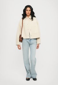 Beige cropped jacket with a button front and patch pocket, paired with light blue flared jeans and black flats, holding a brown shoulder bag.