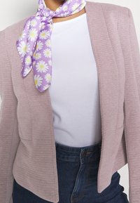 Kvinna iklädd en lavendelfärgad ribbad jacka, vit skjorta, mörkblå jeans och en lila scarf med vita och gula prästkragar knuten runt halsen.