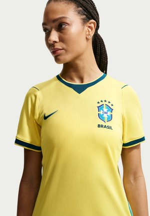 Femeie cu părul împletit purtând un tricou galben de fotbal al Braziliei, cu detalii verzi și emblema CBF pe piept, privind spre o parte.