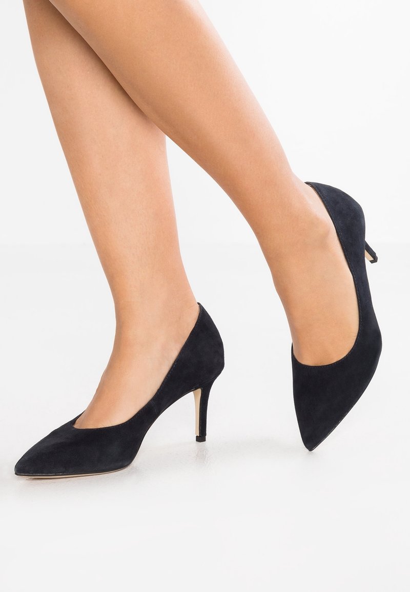 Escarpins en daim noir à talons hauts, bout pointu et talon fin. Les chaussures présentent une texture lisse et une découpe arrondie autour de la cheville.