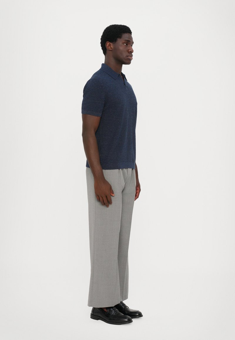 Polo shirt navy a maniche corte con un motivo strutturato, abbinato a pantaloni larghi grigio chiaro e scarpe nere senza lacci. Sfondo neutro.