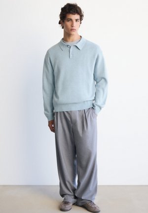 Jeune homme aux cheveux bouclés portant un pull polo bleu clair à manches longues, un pantalon gris ample et des baskets grises, debout devant un mur uni.