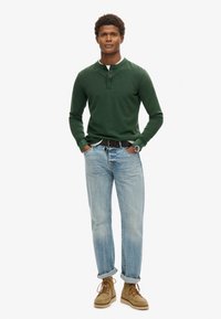 Camisa henley de manga larga verde con botones, combinada con jeans de mezclilla azul claro y botas de cordones beige. Diseño sencillo y corte relajado.