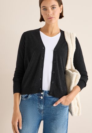 Cardigan nero a maglia con scollatura a V e chiusura a bottoni, abbinato a una maglietta bianca e jeans azzurri chiari. Mani nelle tasche, borsa a spalla appesa.