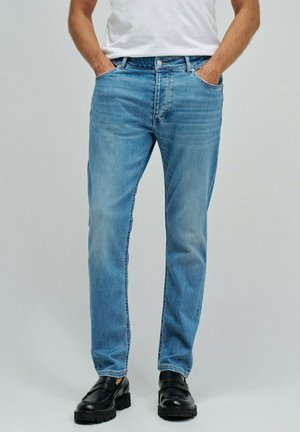 Hombre con pantalones vaqueros rectos de color azul claro, con las manos en los bolsillos y zapatos de cuero negros, de pie frente a un fondo gris liso.