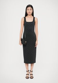 Schwarzes Strickkleid mit quadratischem Ausschnitt, das ein durchgängiges Logo-Muster zeigt, kombiniert mit einer kleinen schwarzen Umhängetasche und schwarzen Riemchensandalen.