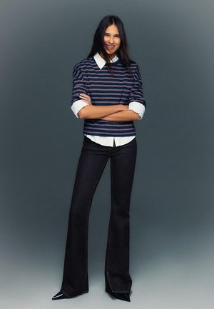 Pulover cu dungi navy și mansete albe, asortat cu blugi evazați închiși la culoare. Ținuta este completată cu pantofi cu toc subțire, negri. Fundalul este gri.