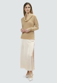 Beige gestrickter Pullover mit Kapuze, kombiniert mit einem langen, cremefarbenen Satinrock mit seitlichen Schlitzen und weißen High Heels.