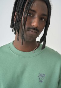 Green cotton sweatshirt with a crew neckline and ribbed cuffs. Features a purple embroidered bird design on the left chest.