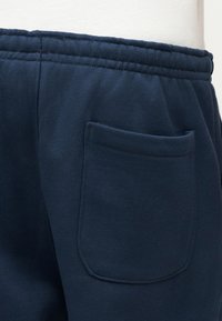 Κοντινή όψη της μέσης και της πίσω τσέπης με ραφές σε σκούρο μπλε φόρμα, πάνω σε ανοιχτό μανεκέν.