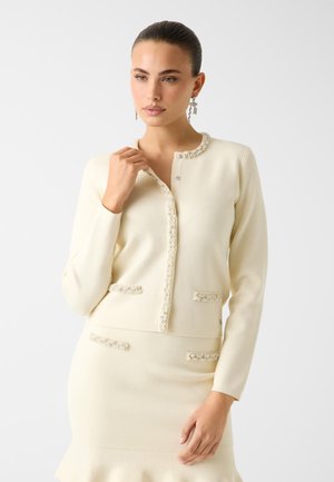 Crèmekleurig gebreid vest met een ronde hals, parelaccenten op de voorkant en twee textuurzakken. Gladde stoftextuur.