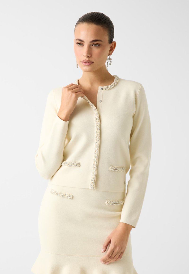 Cremefarbene Strickjacke mit rundem Ausschnitt, Perlendetails auf der vorderen Blende und zwei strukturierten Taschen. Glatte Stofftextur.