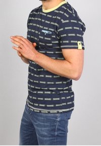 Mies, ktorý má na sebe modrú pásikovanú košeľu s krátkym rukávom a modré džínsy, stojí s rukami zloženými pred sebou a hľadí doprava na jednoduchom svetlom pozadí.