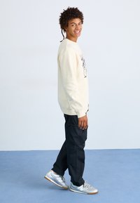 Sudadera beige clara con estampado gráfico, pantalones cargo negros y zapatillas plateadas. El modelo está de pie y sonriente frente a un fondo liso.