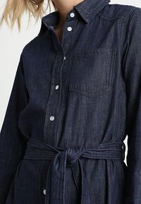 Mörk denimskjortklänning med knappstängning fram, spetskrage, långa ärmar, bröstficka och knytrem detail vid midjan.