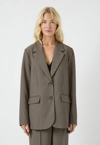 Bruine op maat gemaakte blazer met een gestructureerde revers, twee voorkleurige zakken, twee knopen en een gladde textuur. Draagt over een zwarte top.