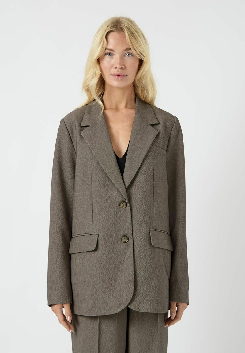 Bruine op maat gemaakte blazer met een gestructureerde revers, twee voorkleurige zakken, twee knopen en een gladde textuur. Draagt over een zwarte top.