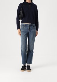 Person, der einen marineblauen Pullover, blaue Jeans, schwarze Sneaker trägt und eine schwarze Umhängetasche hält, steht vor einem weißen Hintergrund.