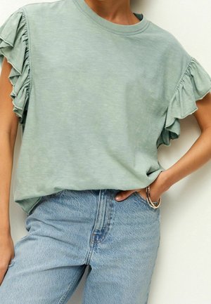 Helles grünes kurzärmeliges Oberteil mit Rüschendetails an den Schultern, kombiniert mit hochtaillierten hellblauen Jeans. Weicher Stoff, lässige Passform.