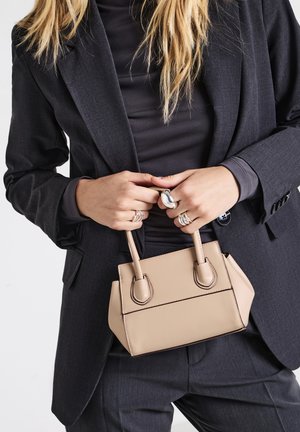 Person in dark suit holding beige handbag with both hands, wearing multiple silver rings, and wearing a black top underneath the blazer.