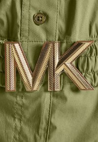 Boucle de logo MK métallique doré sur une ceinture en tissu vert olive avec des coutures visibles et un bouton au-dessus de la boucle.