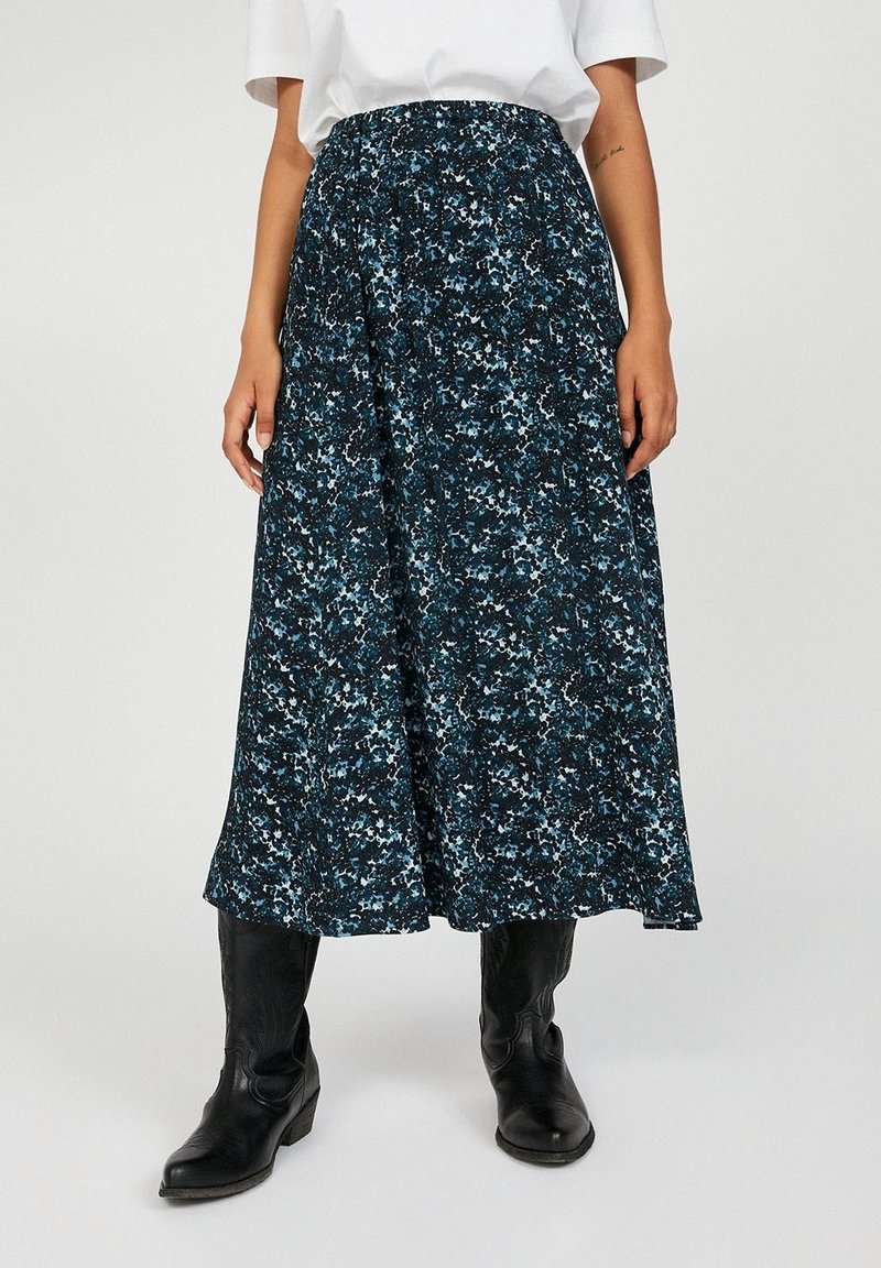 Femme portant une jupe mi-longue bleue et noire à fleurs avec un haut blanc et des bottes de cowboy noires, debout devant un fond uni.