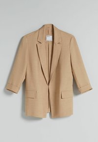 Blazer beige en tissu texturé, avec un col cranté, des manches trois-quarts et deux poches avant. Design à ouverture frontale.